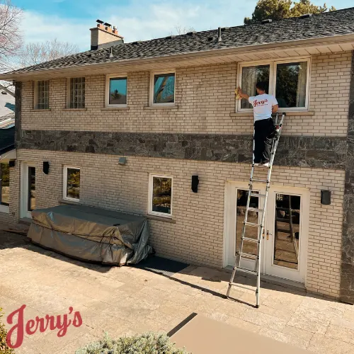A crew member on a ladder cleaning a second-floor window on a yellow-brick house
